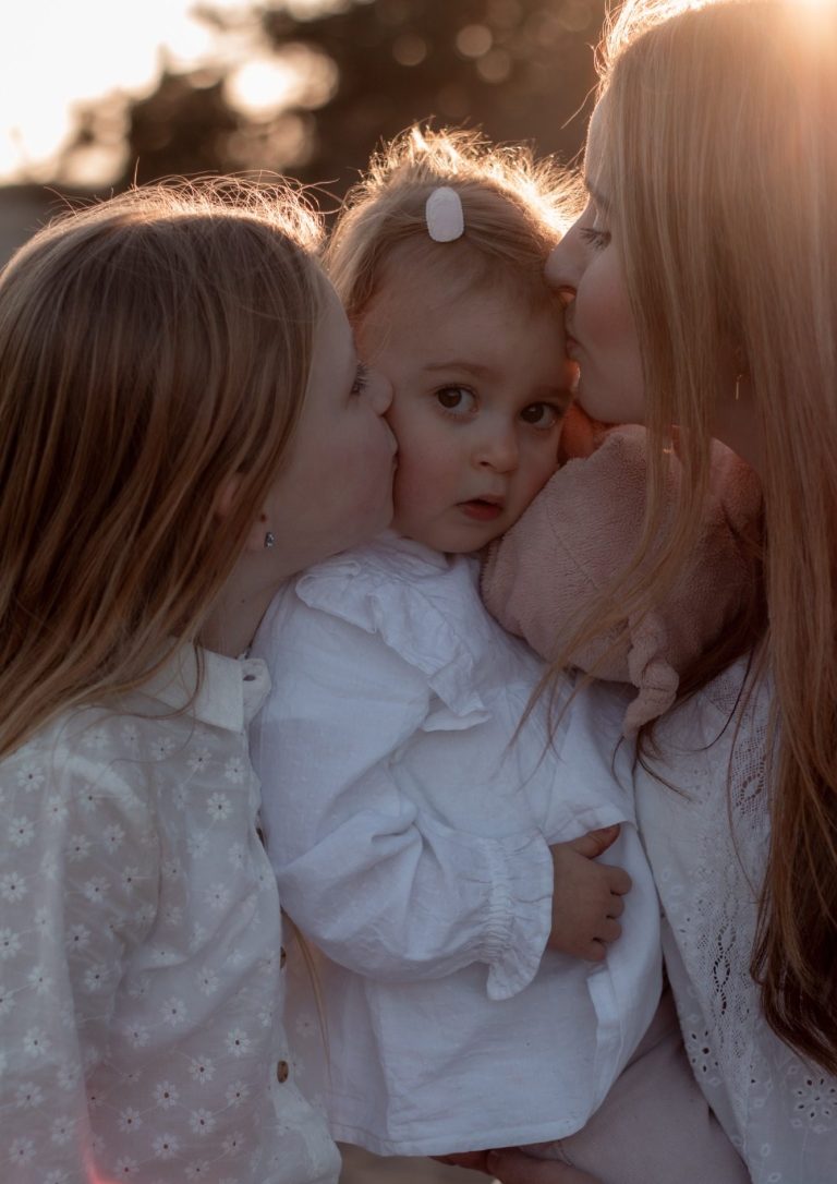 Moeder kust haar kindje terwijl een oudere zus haar op de wang kust, zonsopgang achtergrond.
