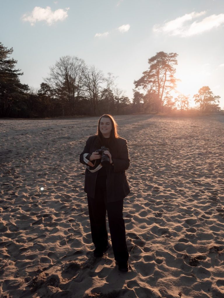 Vrouw met blond haar zit op een bank, werkt op laptop en drinkt uit een glas.