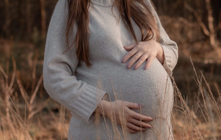 Zwangere vrouw in een warme trui met handen op haar buik, omringd door natuur.