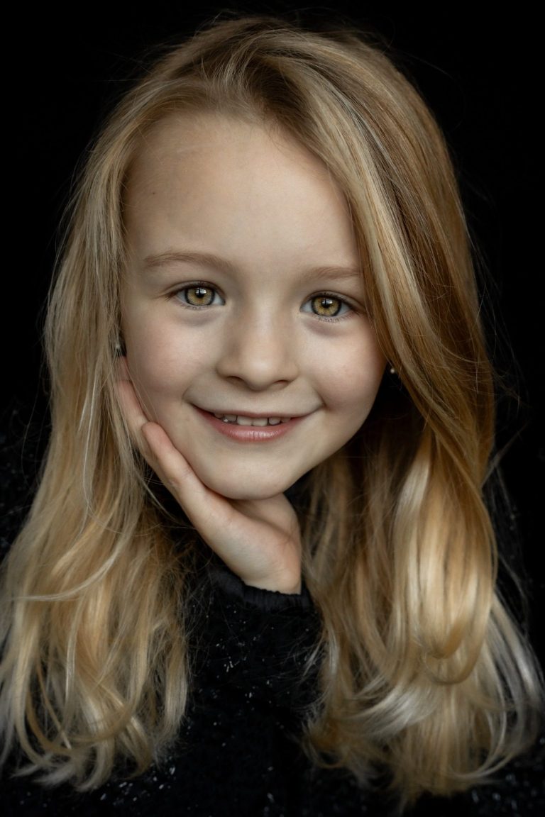 Een glimlachend meisje met lang, golvend blond haar op een donkere achtergrond.