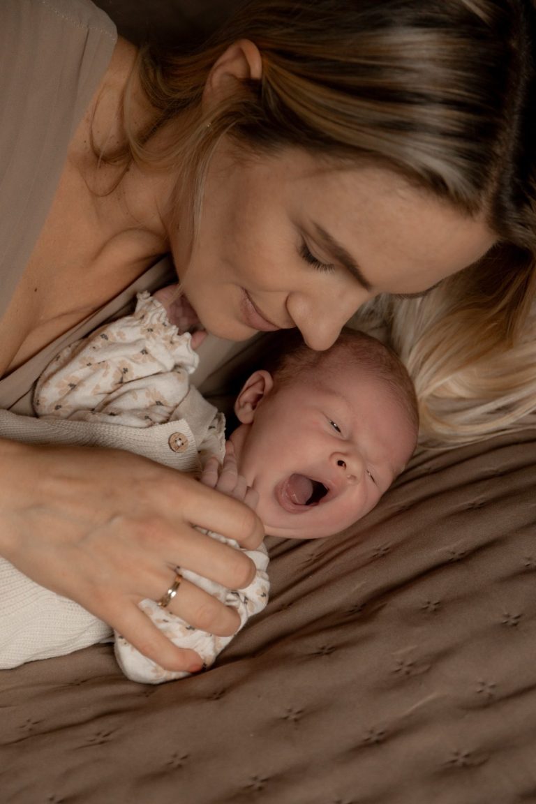 Moeder knuffelt een gapende baby op een bed met zachte, neutrale kleuren.