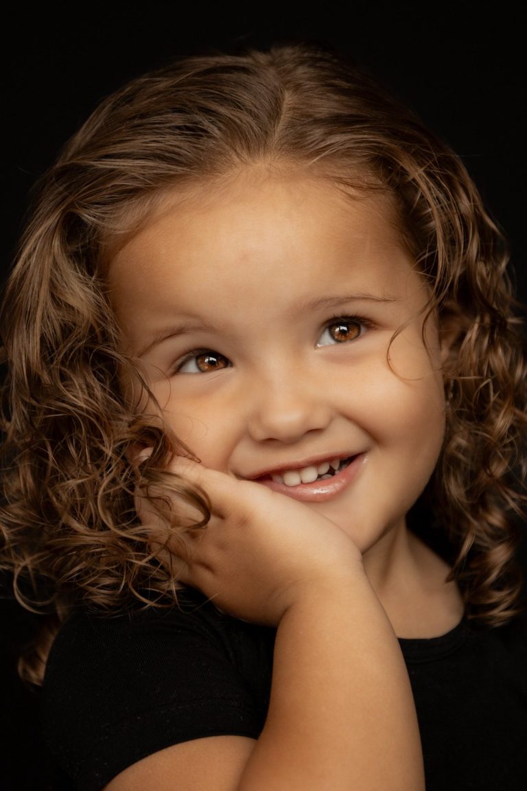 Lachend meisje met krullend haar en een hand op haar gezicht.