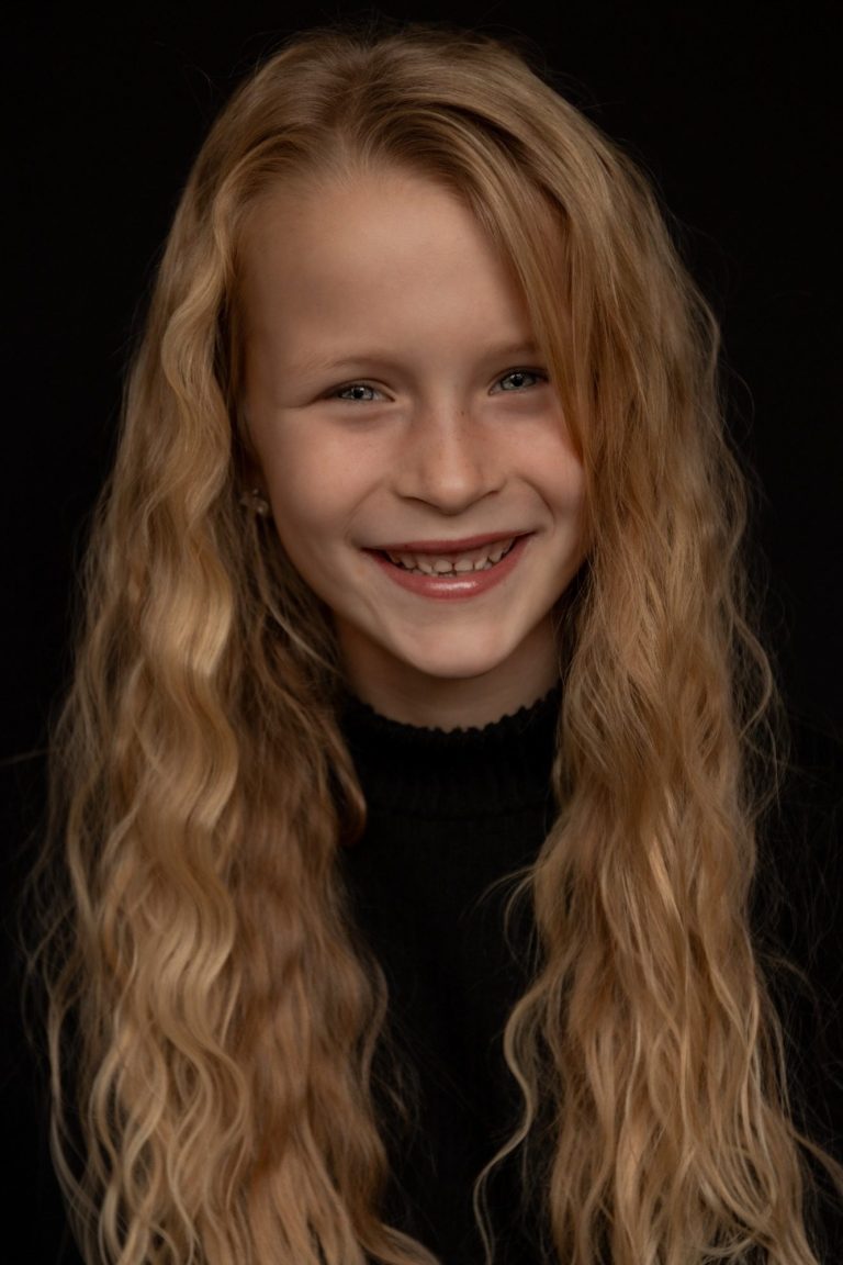 Blonde jongen met krullend haar en een brede glimlach, op een donkere achtergrond.