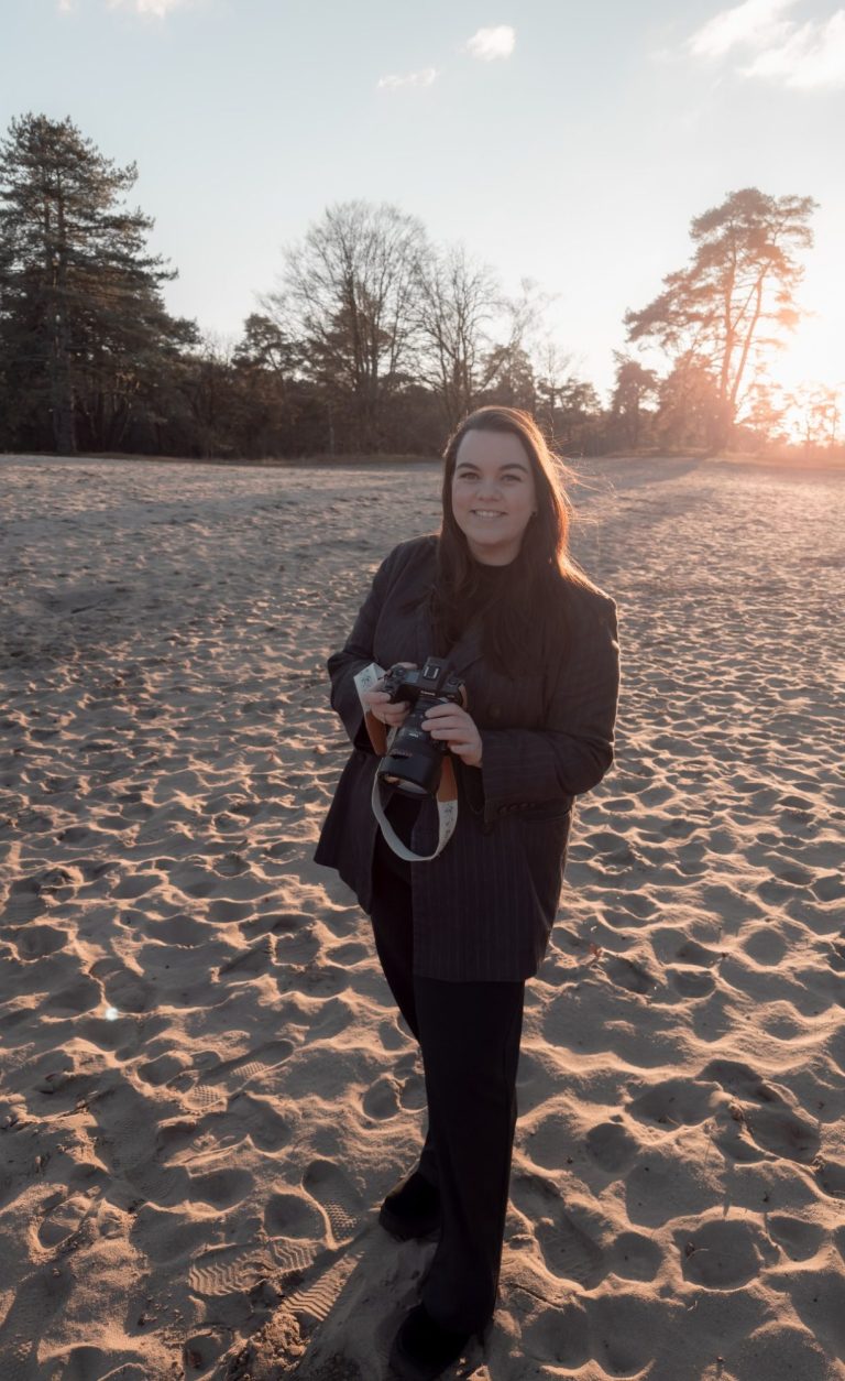 Een vrouw in een beige jas fotografeert buiten bij zonsondergang op een zandachtige ondergrond. Tiffany Kappelle, fotograaf, zacht licht 