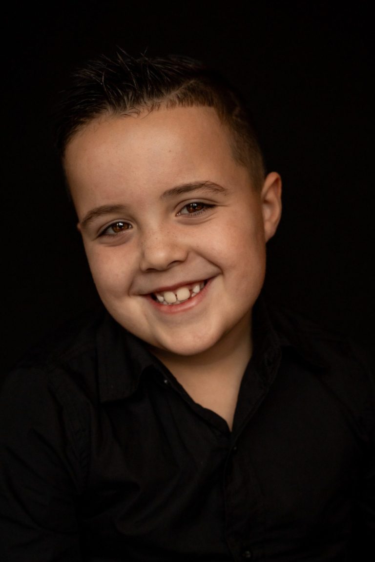 Jongen met een glimlach, gekleed in zwart, op een donkere achtergrond.