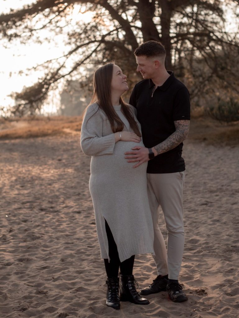 Zwangere vrouw en man staan op het strand en kijken elkaar liefdevol aan.