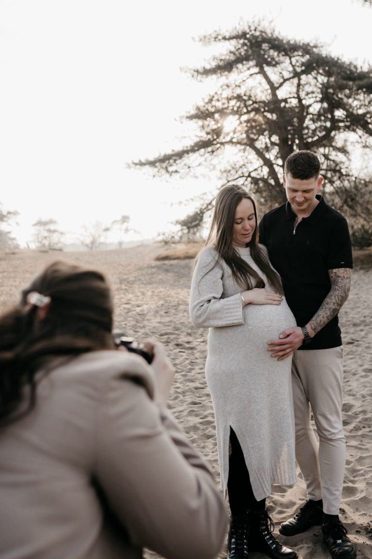Zwangere vrouw poseert met haar partner op het strand, terwijl iemand hen fotografeert.