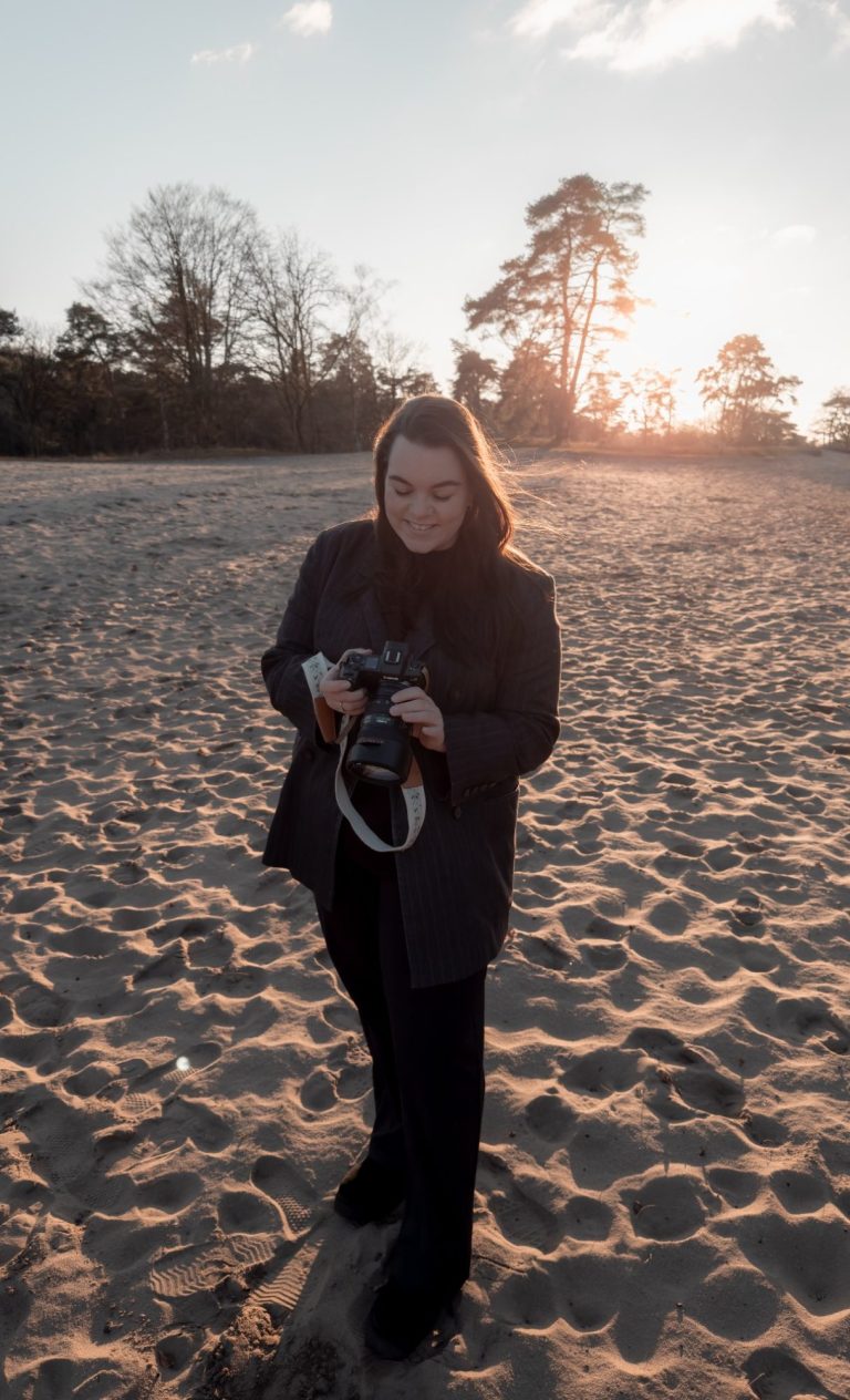 Vrouw in beige jasje houdt een camera vast op een strand bij zonsondergang.