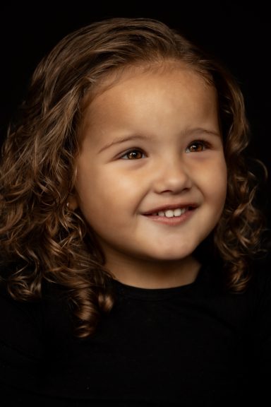 Een lachend meisje met krullend haar in een donkere omgeving.