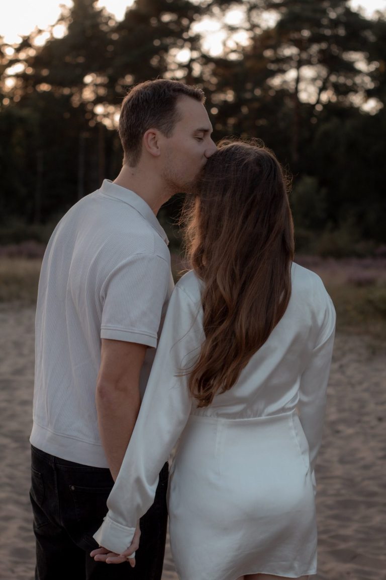 Een stel staat hand in hand, de man kust de vrouw zacht op het voorhoofd.