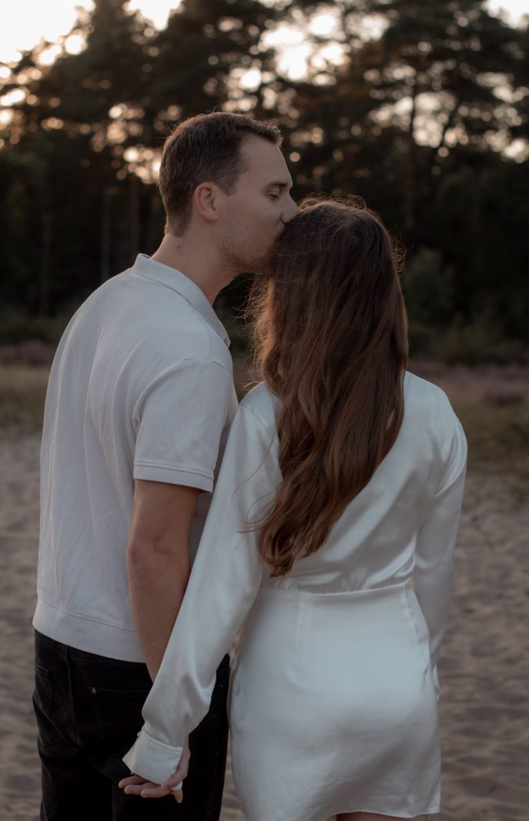 Een stel staat samen op het strand, de man kust de vrouw op het hoofd.