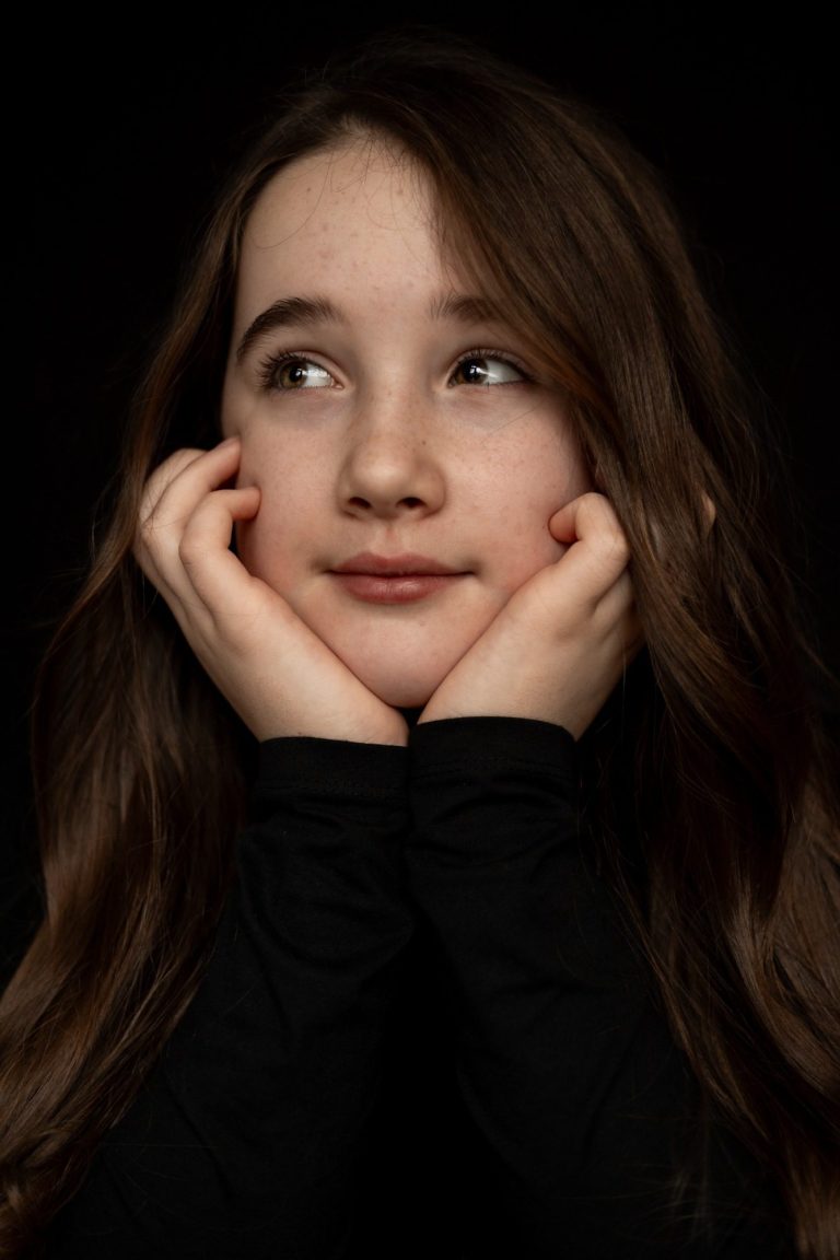 Meisje met lange, bruine haren, met een contemplatieve blik, handen onder kin.