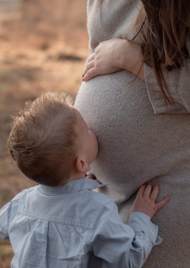Een peuter kust een zwangere buik, terwijl hij met zijn handen de buik vasthoudt.