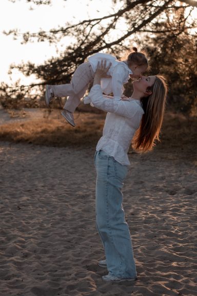 Een vrouw tilt een kind op terwijl ze in de lucht kijkt, op een zonnige zandige locatie.