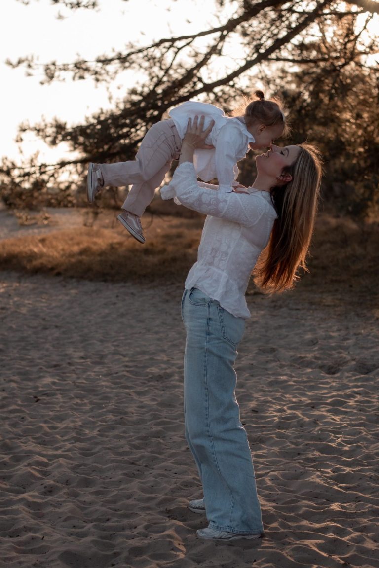 Moeder tilt lachend haar kind op in een zonnige, natuurachtige omgeving.