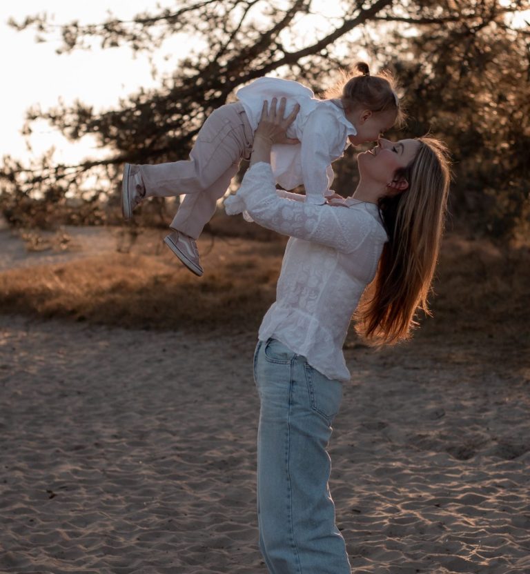 Vrouw tilt lachend een peuter op in een natuurlijke omgeving tijdens zonsondergang.