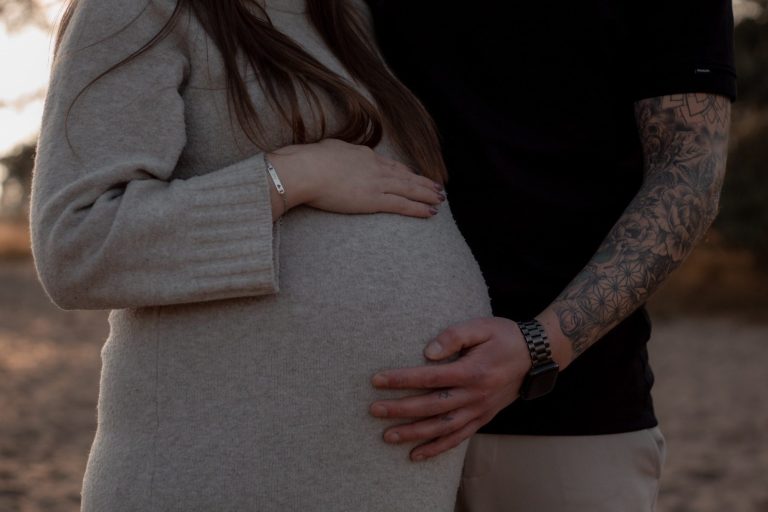 Een zwangere vrouw met een hand op haar buik, omarmd door een man.