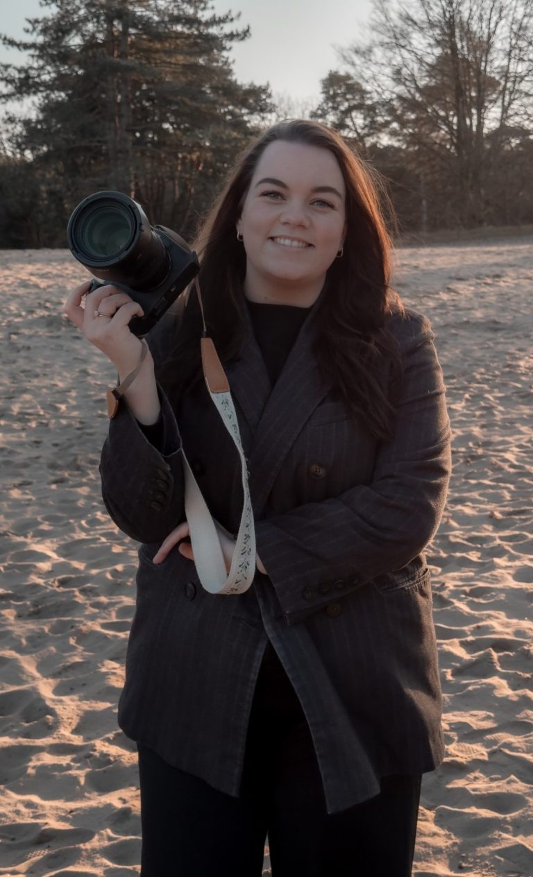 Vrouw in beige jasje houdt een camera vast op een strand bij zonsondergang.