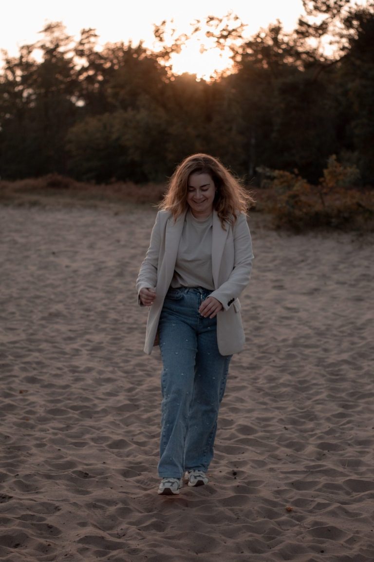 Een vrouw met krullend haar wandelt op een zandige ondergrond tijdens zonsondergang.