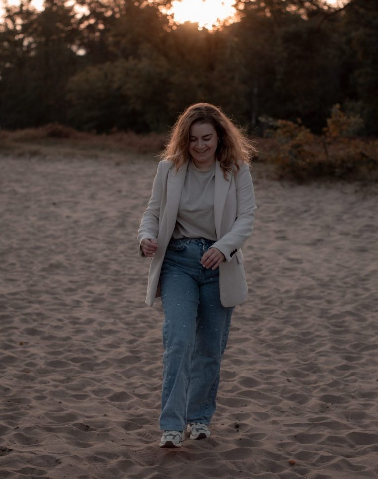 Vrouw met krullend haar, in een licht jasje, wandelt over een zandige ondergrond.