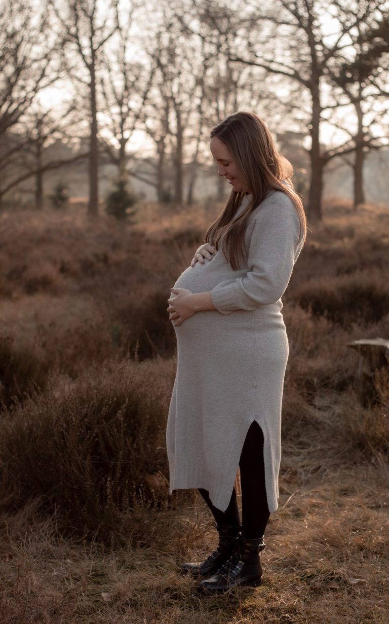 Zwangere vrouw in een lichtgrijze jurk staand in een natuurlijke omgeving.