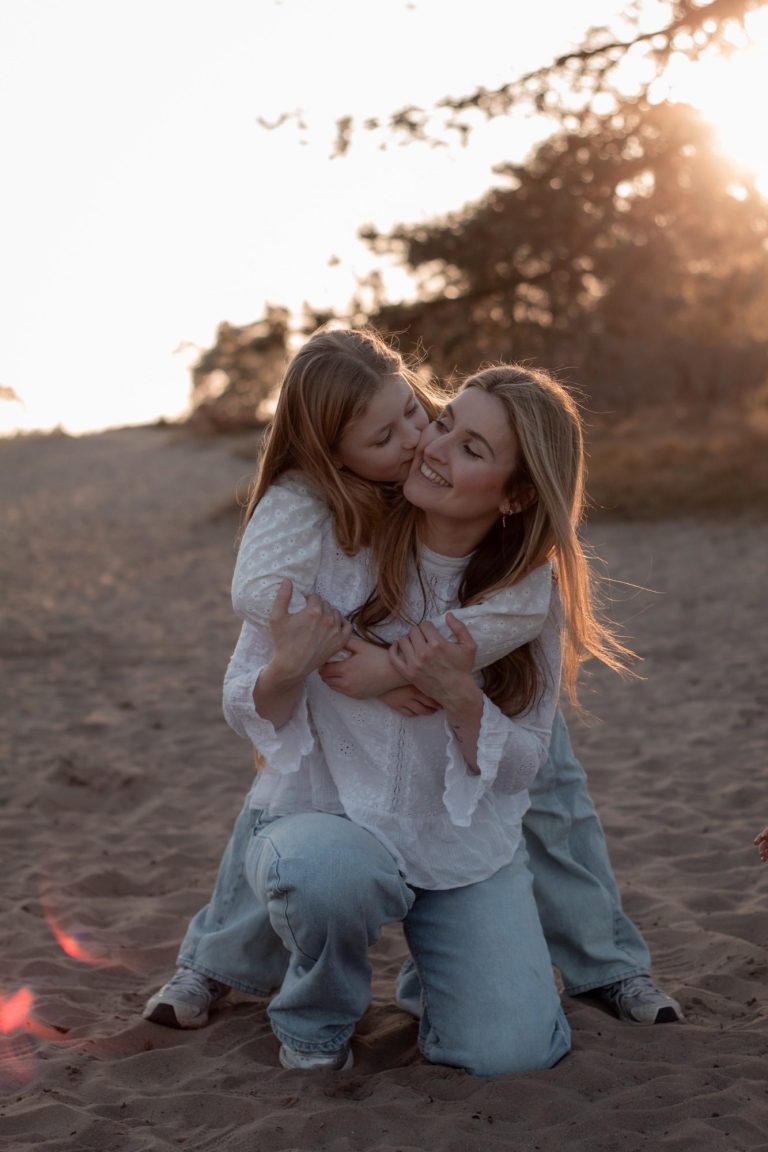 Vrouw en meisje omhelzen elkaar op een zandachtige plek bij zonsondergang.