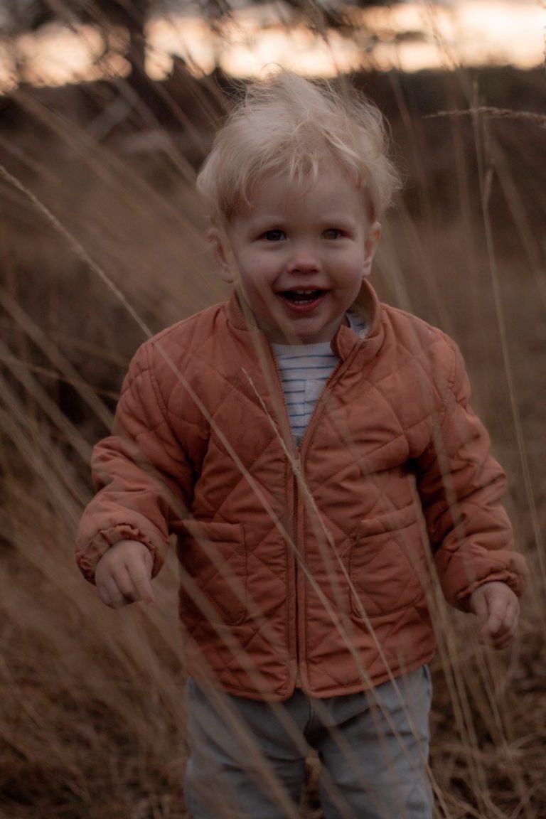 Blonde peuter in een oranje jasje staat lachend tussen het gras.